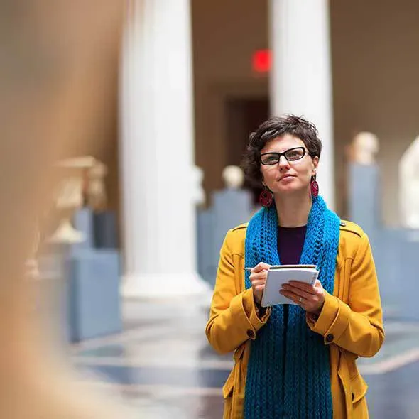 Woman with glasses in yellow coat and blue scarf holding a notebook and pen looking at art in a gallery