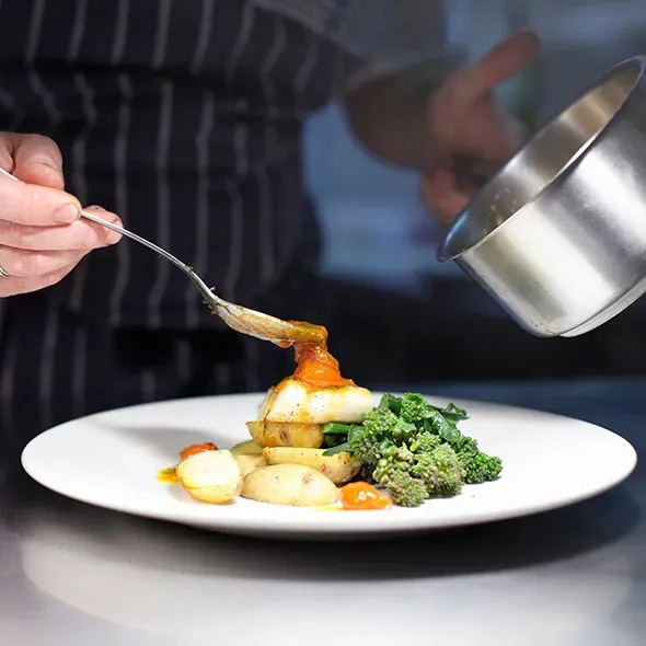 A chef spooning over a sauce on to a dish of food in a professional kitchen