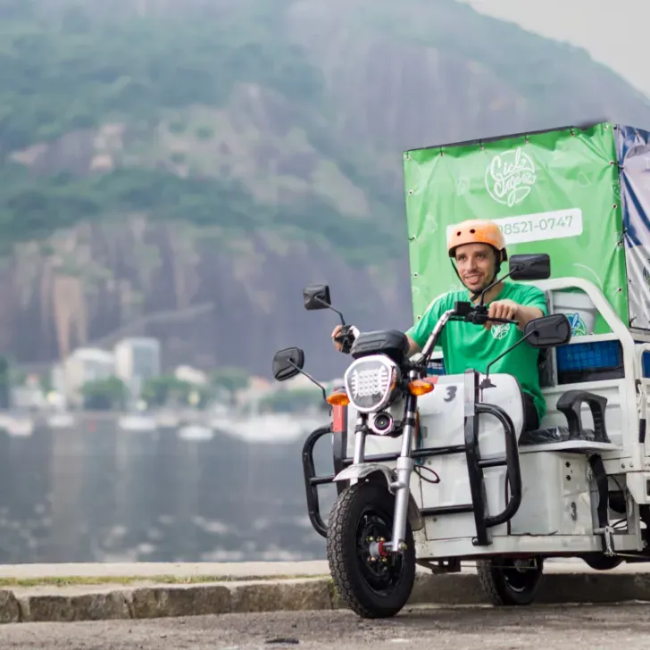 Man on yellow motorised tricycle wearing a green shirt and yellow helmet riding past a river with green cliff face in background